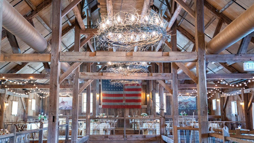 second floor of barn tables around in half circle around balcony opening to first floor with chandeliers lit from wood beamed ceiling